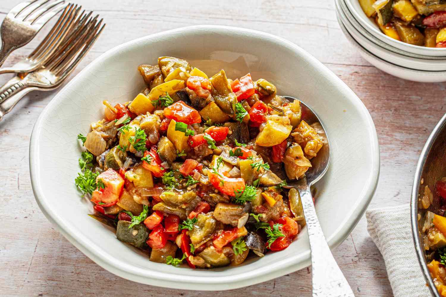 Bowl of Classic Ratatouille with a Spoon Next to More Utensils, Stack of Bowls, and Pan with More Ratatouille 