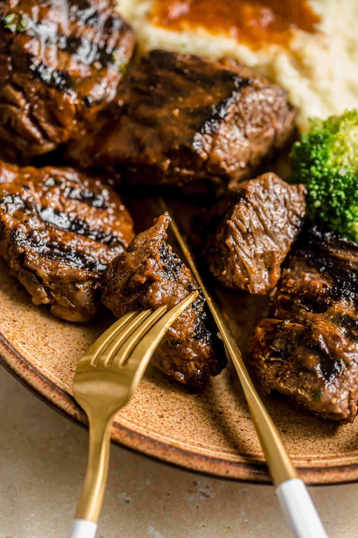 Close-up: fork and knife cutting into a piece of steak tip on a plate wiht mashed potatoes and gravy and oven-roasted broccoli and asparagus on a plate with a fork