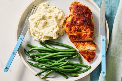 A plate with seasoned cooked chicken breast mashed potatoes and green beans accompanied by a fork and knife