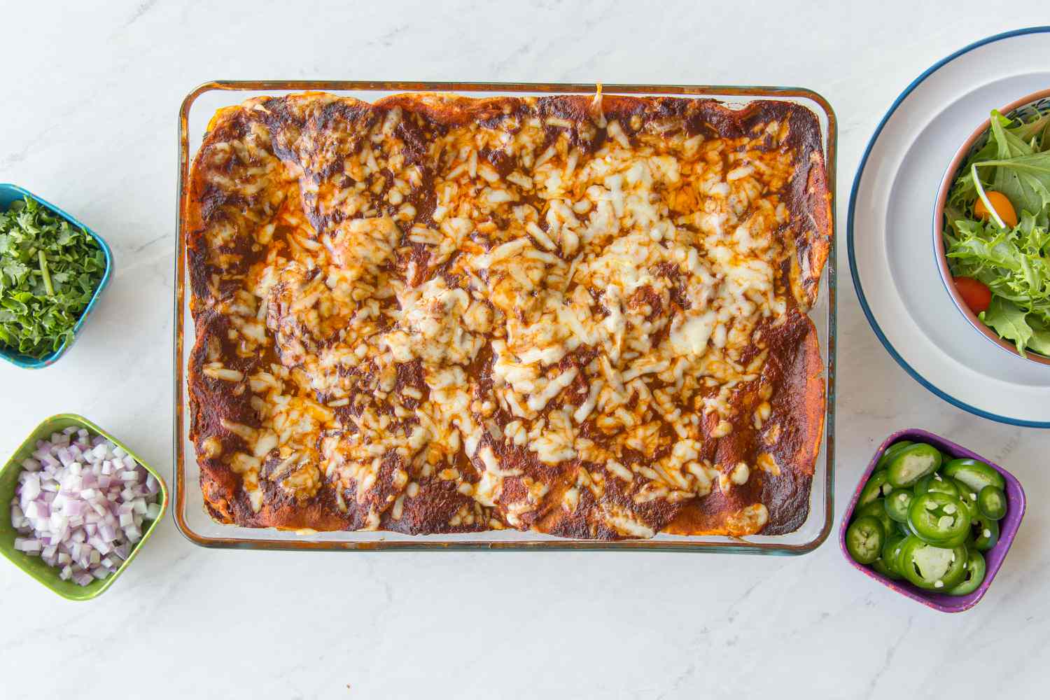 Baked Chicken Enchilada Casserole Surrounded by Bowls of Toppings (Chopped Onions, Sliced Jalapenos, and Cilantro), and a Bowl of Side Salad on a Stack of Plates