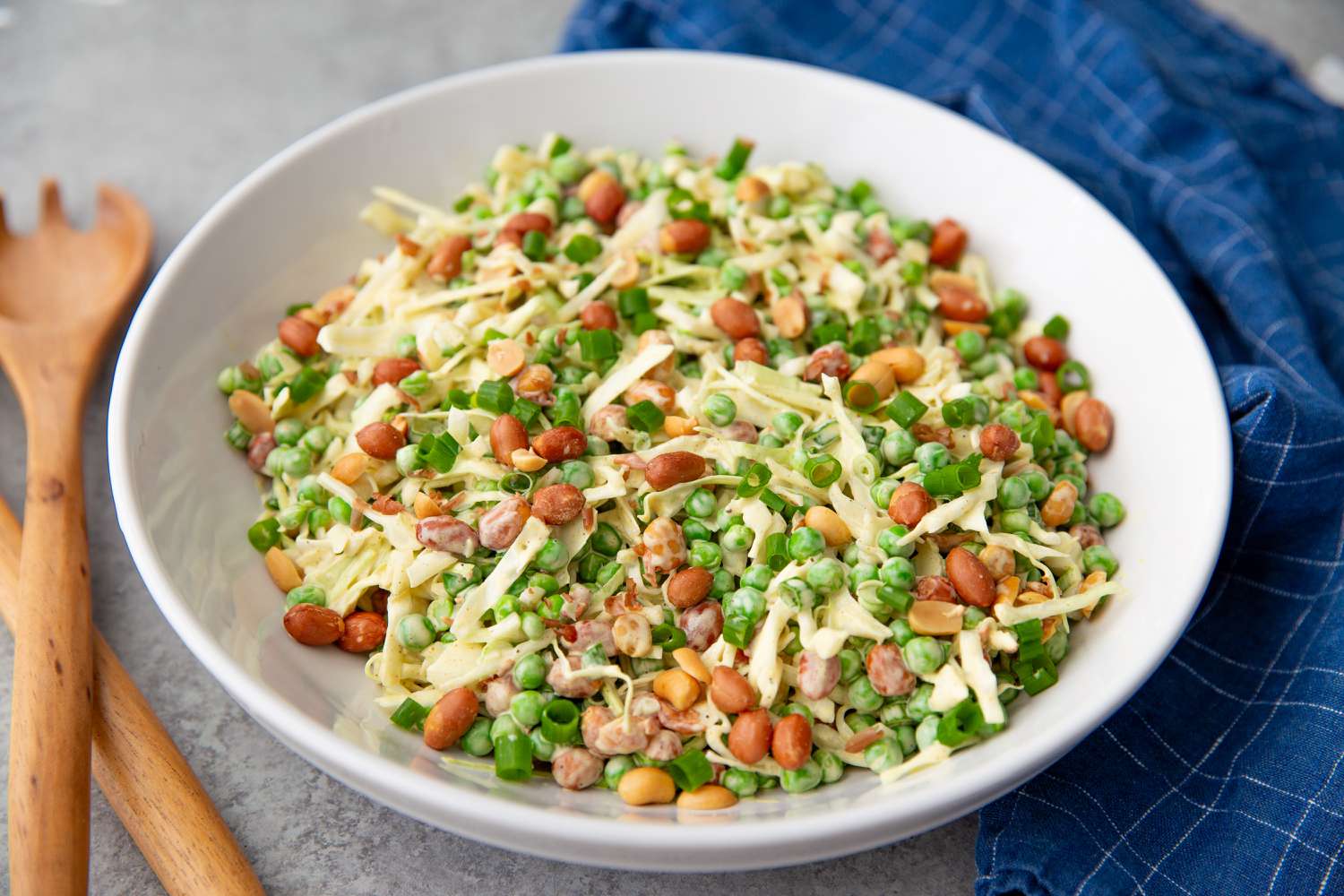 White bowl of cabbage slaw on a gray table with a blue checkered napkin underneath and wooden serving utensils to the side