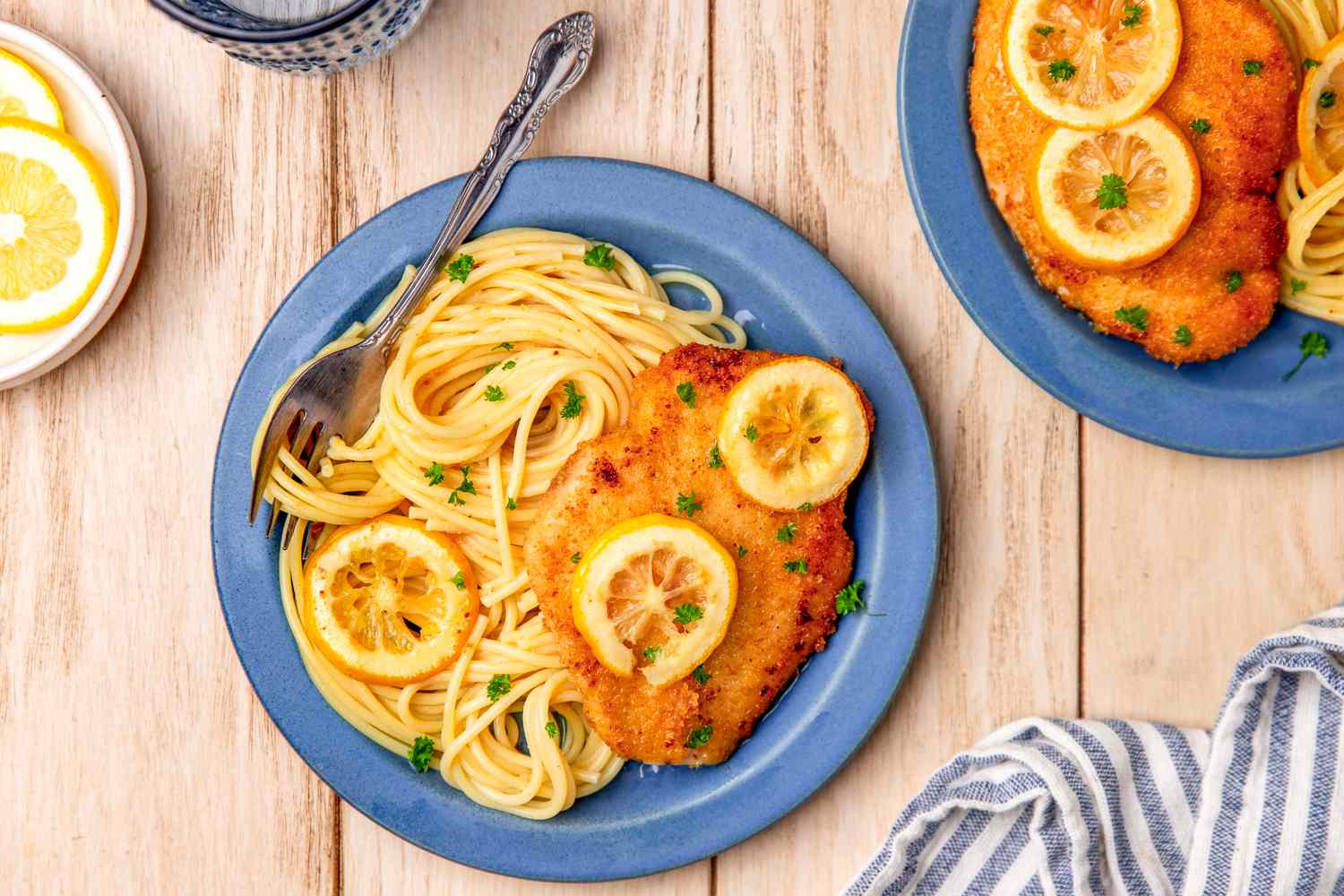 Italian butter chicken (petti di pollo al burro) with pasta on two plates at a tablee setting with a plate of lemon slices, a glass of water, and a table napkin