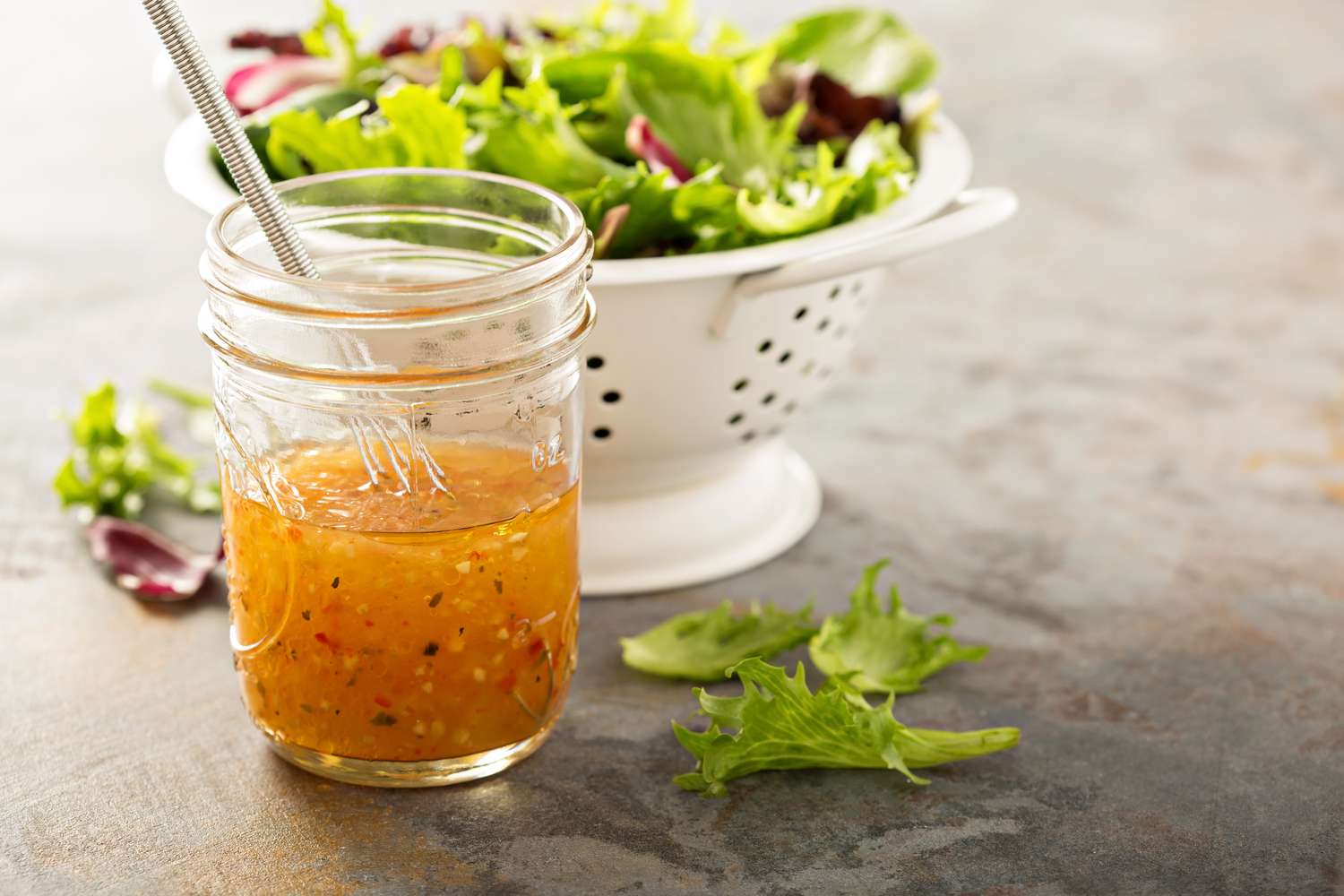 Italian vinaigrette in a mason jar with fresh salad greens in a colander in the background.