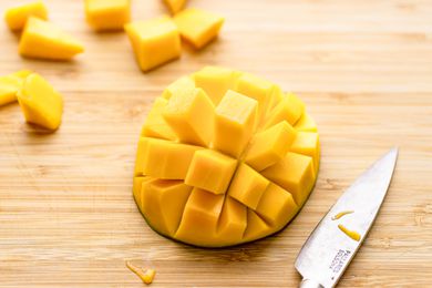 Halved Mango With Cut Using the Cross-wise Cut Method on a Cutting Board Next to a Knife and More Cubed Fruit in the Background