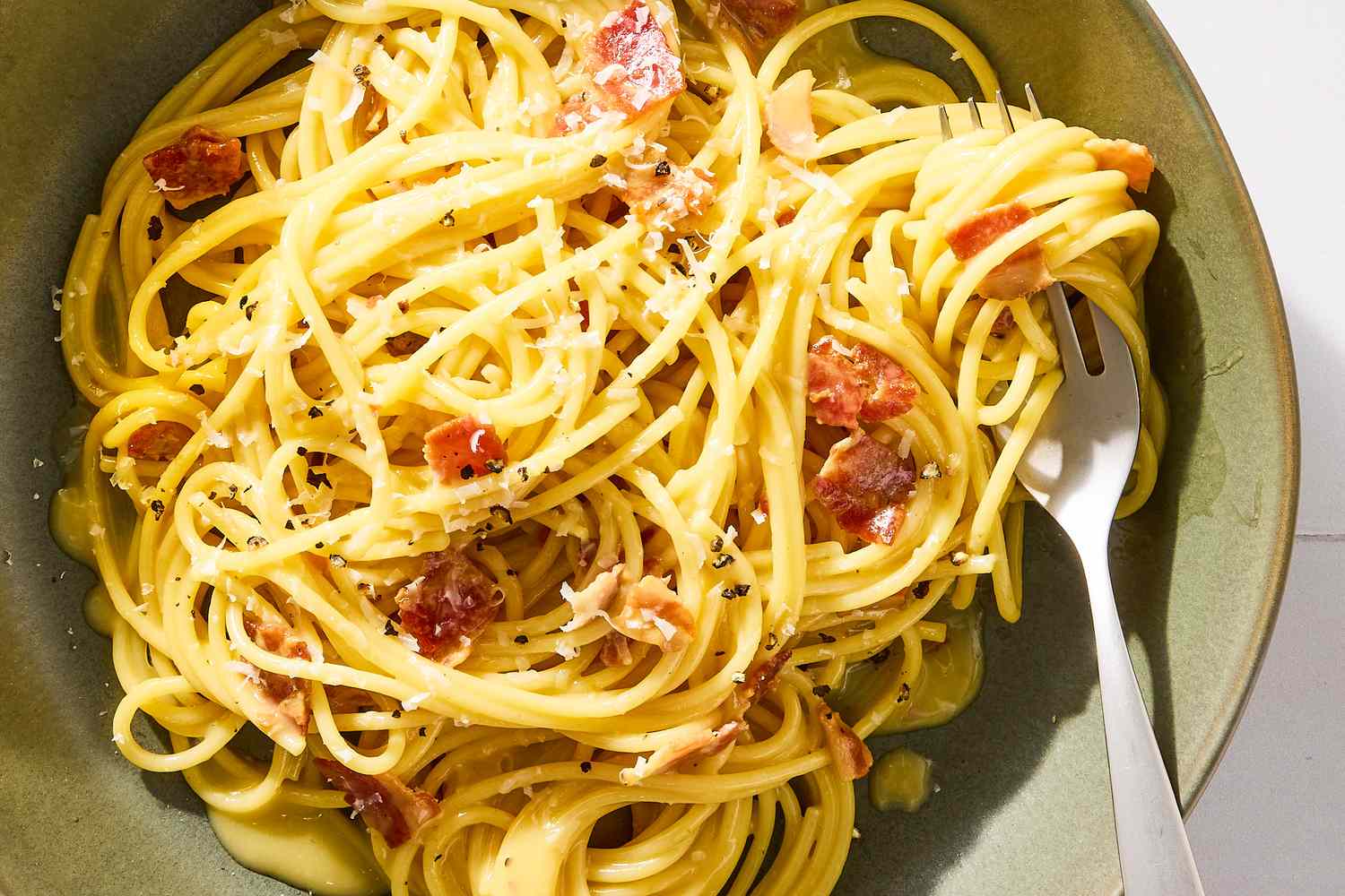 A plate of spaghetti carbonara with grated cheese and bacon bits accompanied by a fork