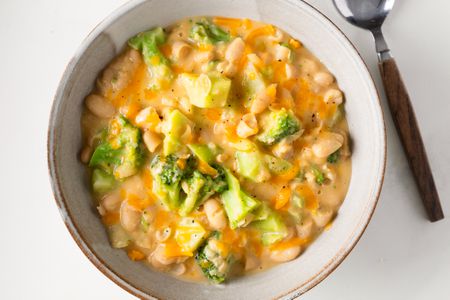 Overhead view of a stoneware bowl of Broccoli Cheddar Creamy Beans next to a spoon and all on a white background
