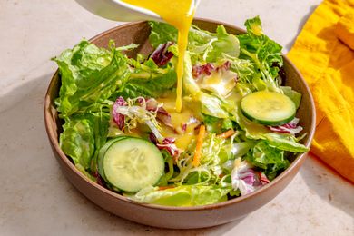 Angled view of pouring Miso-Maple Dressing over lettuce and cucumbers in a brown bowl all on a tan countertop