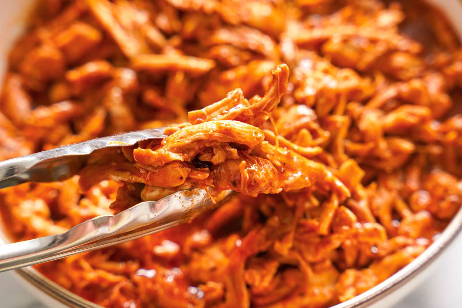 Tongs Holding a Bit of Pulled Chicken and in the Bowl Below It, More Pulled Chicken