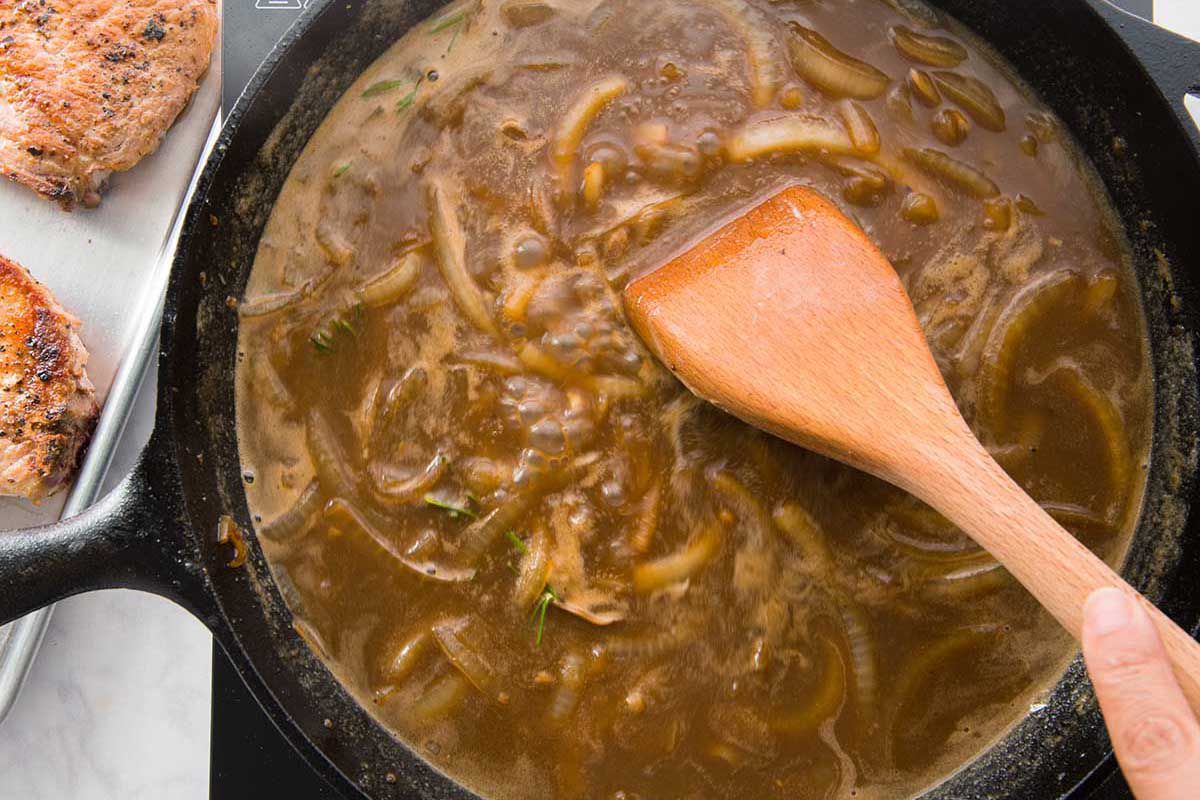 Onions in a cast iron skillet filled with gravy and being stirred with a wooden spoon.