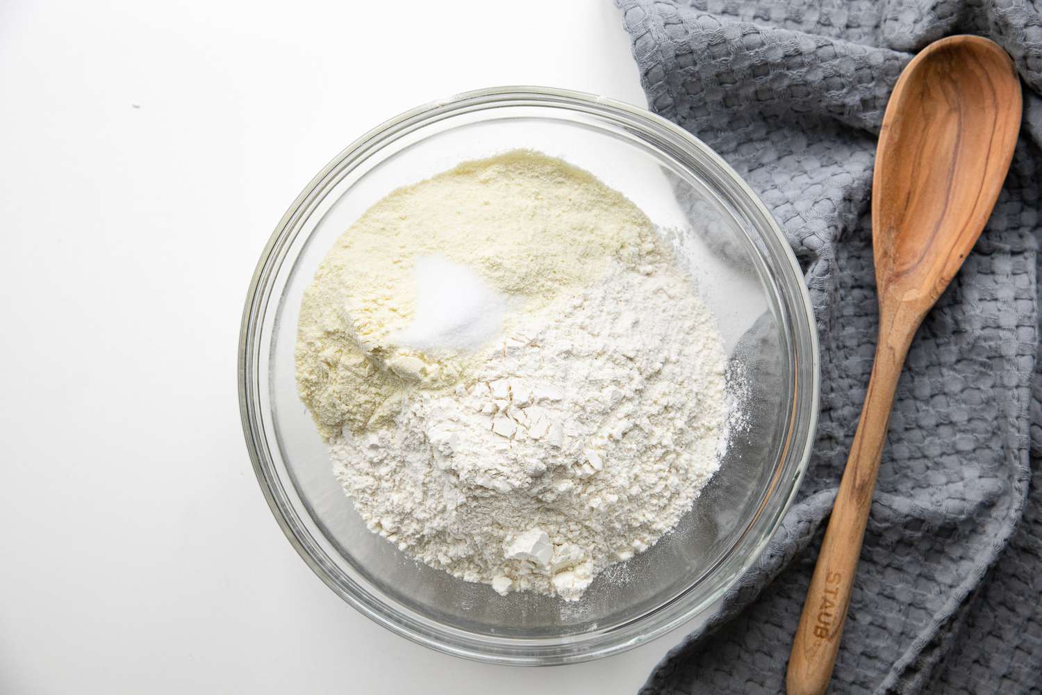 Dry Ingredients (Semolina, Salt, Flour, etc.) Added to a Bowl Next to a Kitchen Towel and a Wooden Spoon for Homemade Spaetzle Recipe
