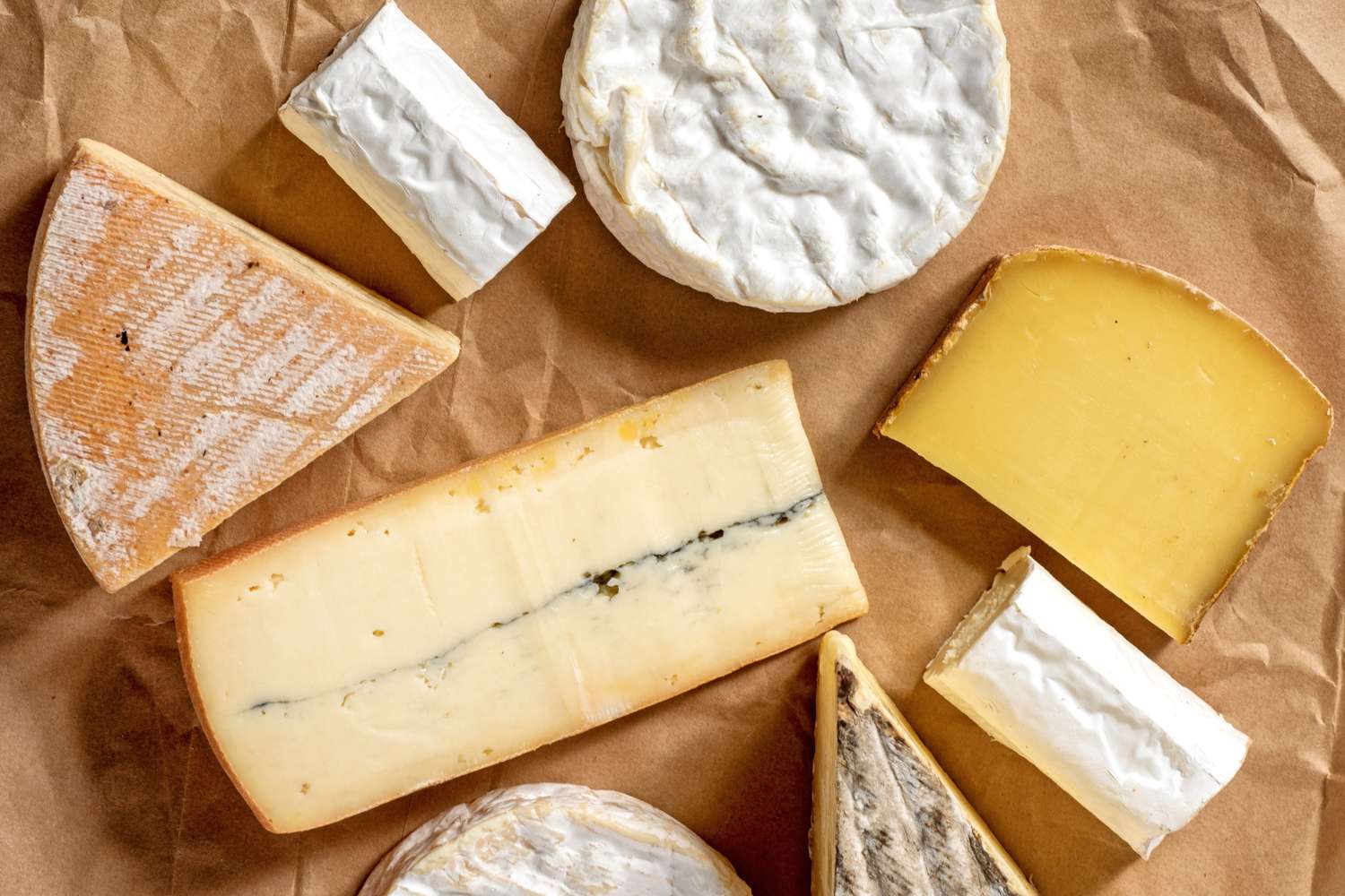 Various types of cheese arranged on brown parchment paper including soft and hard cheese varieties in multiple shapes and textures