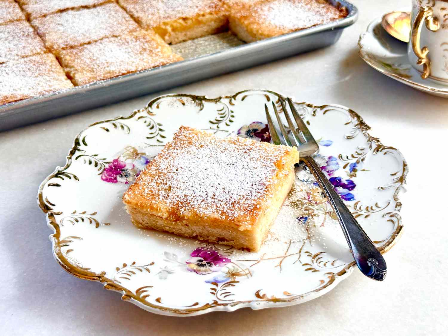 A lemon square on a decorative plate with a fork, in front of a tray of additional lemon squares