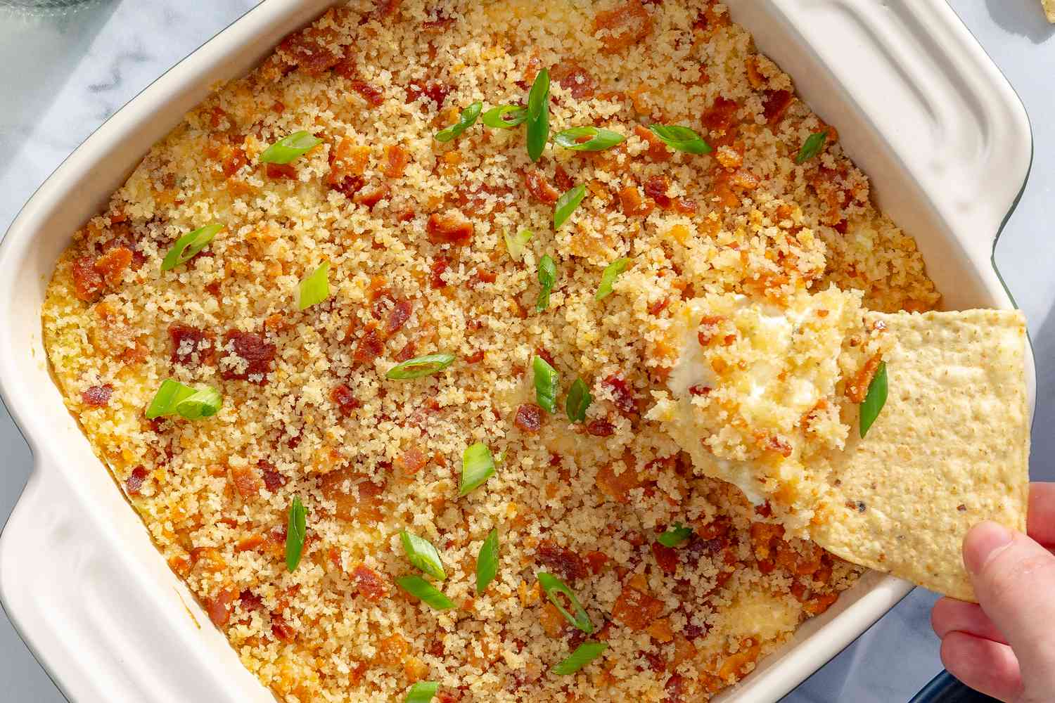 Overhead shot of a white square baking dish with jalapeño popper dip, and a hand holding a tortilla chip scooping some of the dip out