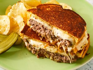 A patty melt sandwich with melted cheese and caramelized onions, served with potato chips and pickle slices on a green plate