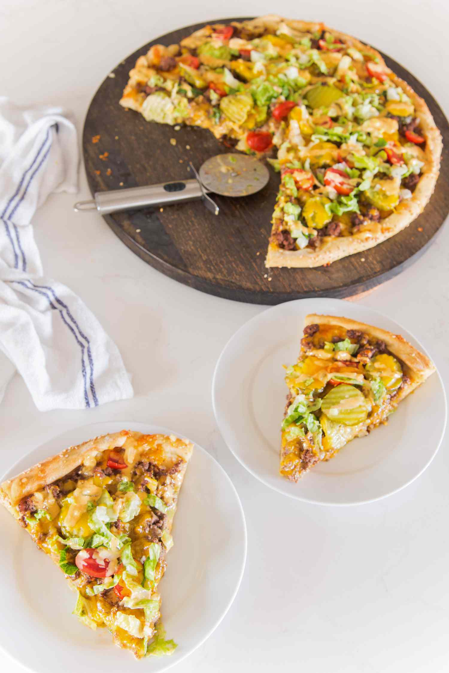 Slice of Cheeseburger Pizza on Small Plate, and in the Background, Another Slice on a Plat and More Pizza on a Circular Wooden Tray, and a Kitchen Towel