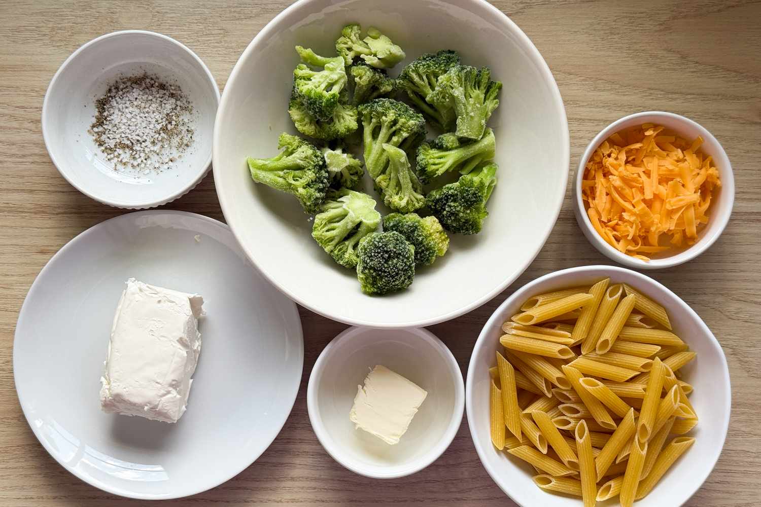 Ingredients for a macaroni and cheese dish including broccoli, pasta, cheddar cheese, cream cheese, butter, and pepper in bowls