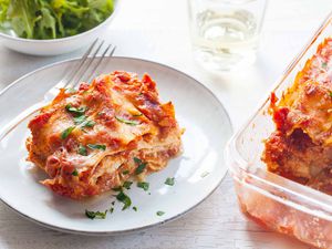 Horizontal view of white plate with slice of small batch lasagna and garnished with chopped parsley. A partial view of the baked lasagna in the glass pan is to the right of the plate. Above the plate to the left is a small bowl of lettuce and a tumbler. A fork is on the left of the plate.