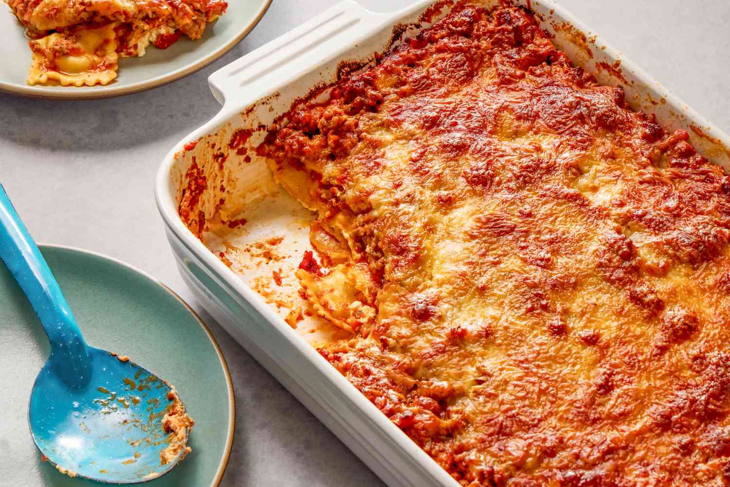 Angled view of a white baking dish of Million Dollar Ravioli Casserole with a serving removed and on a small plate next to a serving spoon on a small plate