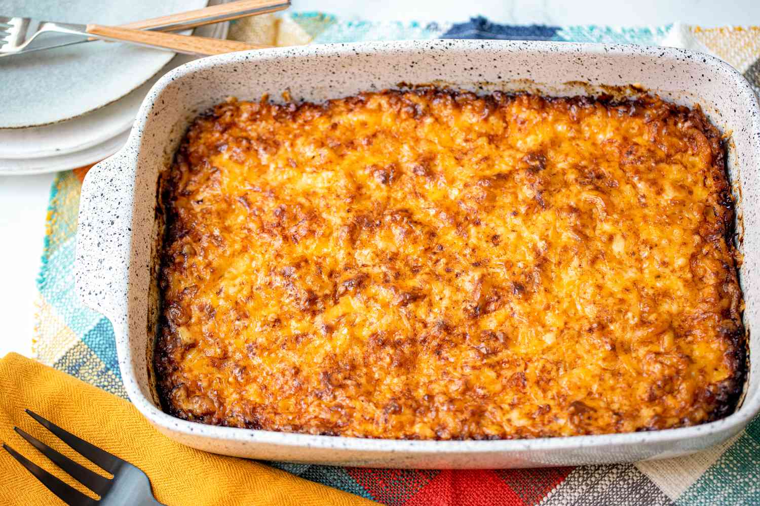 Trinidadian Macaroni Pie in a Casserole Dish on a Placemat Next to a Spatula on a Table Napkin and a Stack of Plates with Utensils on Top