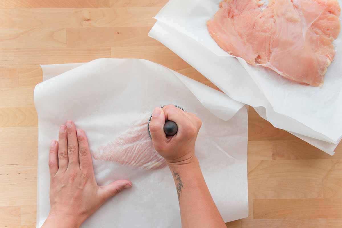 Photo of chicken sandwiched between two sheets of parchment paper and hands are using a meat tenderizer to pound it flat to make the best chicken cordon bleu