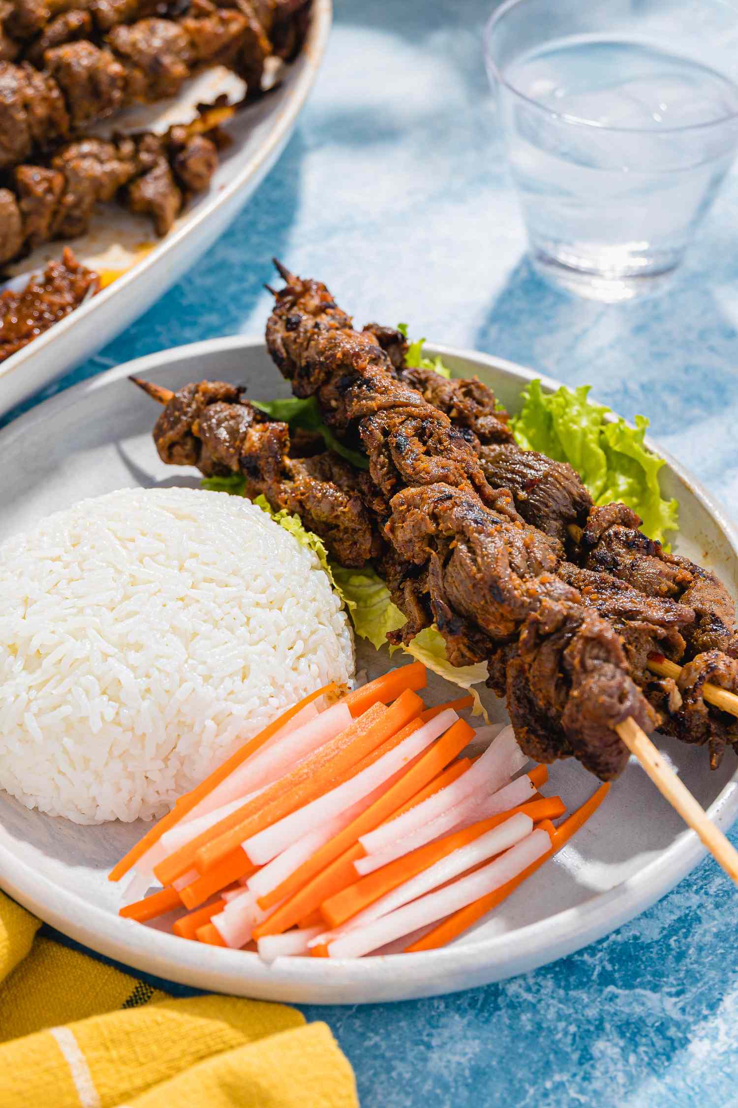 Plate With Sach Ko Jakak (Cambodian Grilled Lemongrass Beef Skewers) With a Mound of Rice and Sliced Daikon Radish and Carrot Pickle, and in the Surroundings, a Glass of Water and a Plate With More Skewers