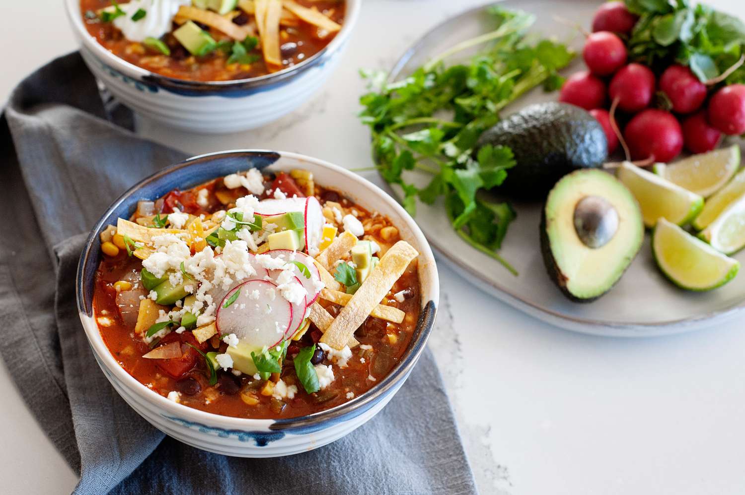 Two bowls of meatless tortilla soup garnished with radishes, cheese, and sour cream and a plate of garnishes next to it.