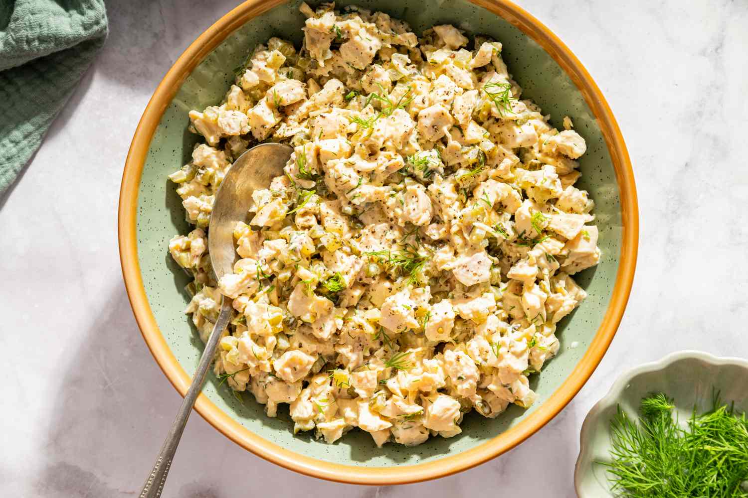 A bowl of chicken salad with pickles and herbs served with a spoon in the bowl on a white marble surface