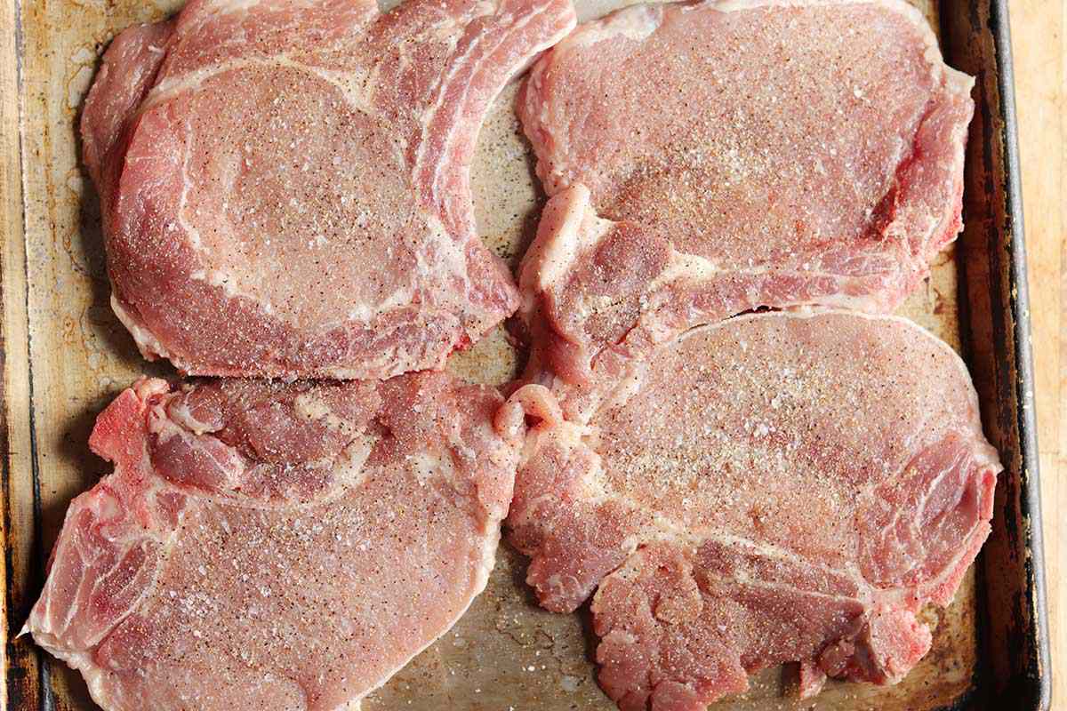 Four raw bone-in pork chops covered in seasoning to show how to cook pork chops.