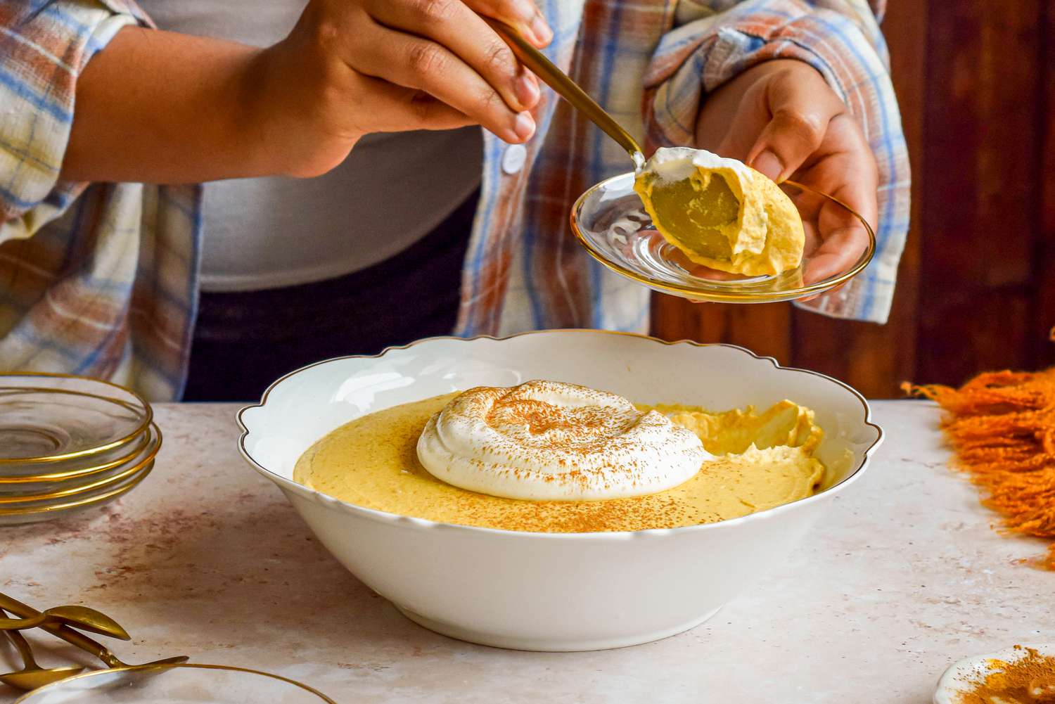 Someone serving a spoonful of 2-ingredient pumpkin mousse onto a small plate at a dinner table. On the dinner table, a bowl with more mousse topped with whipped cream and a sprinkle of pumpkin spice and more small plates with spoons