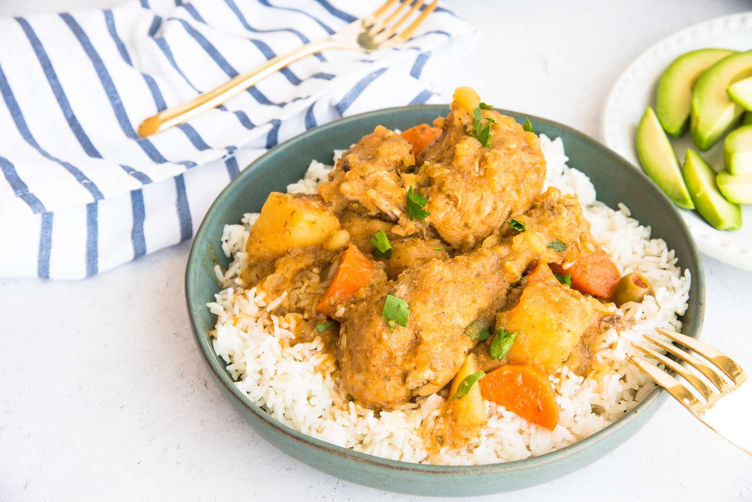 Bowl of Pollo Guisado Served Over Rice Next to a Plate with Sliced Avocado Pieces 