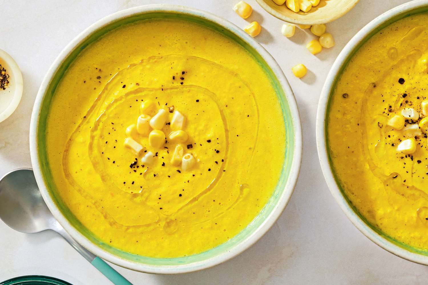Two bowls of corn gazpacho garnished with corn kernels and black pepper with a spoon beside one bowl