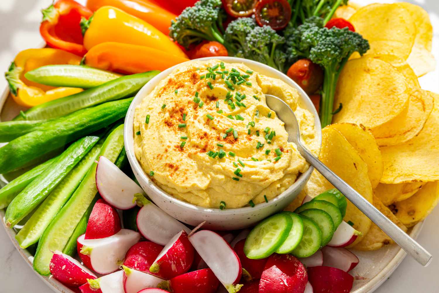 Closeup view of a platter with vegetables and potato chips arranged around a bowl of deviled egg dip all on a white background