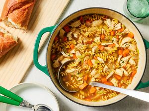A pot of turkey soup with rotini pasta next to a cutting board with sliced baguette
