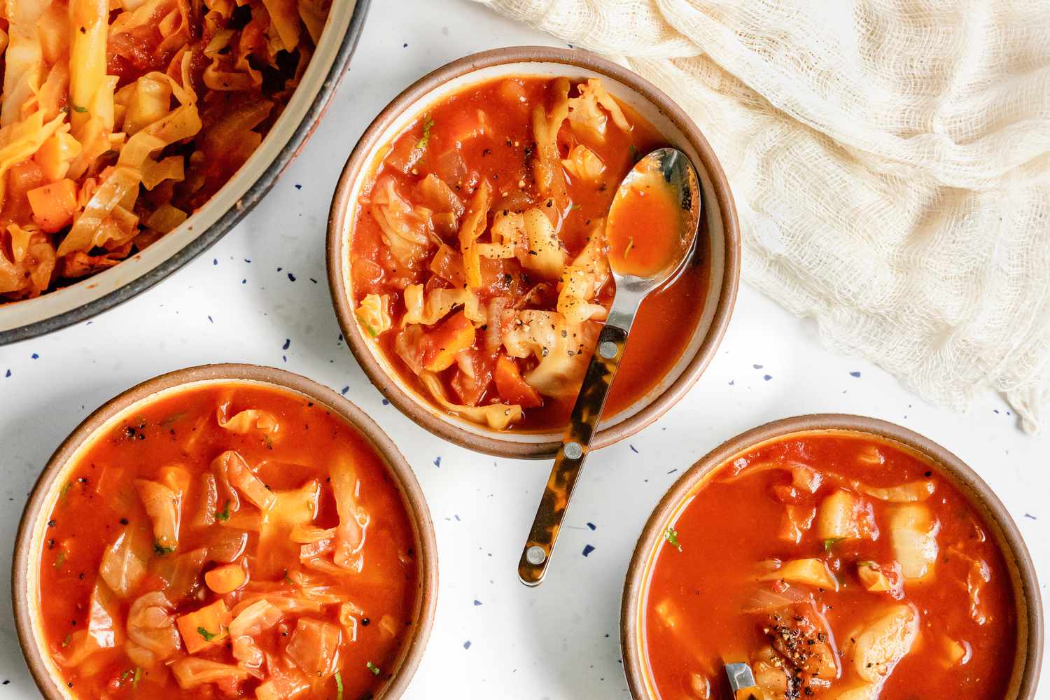 Sweet and Sour Cabbage Soup in Three Bowls with Spoons, and Next to It, a Large Pot with More Soup and a Kitchen Linen