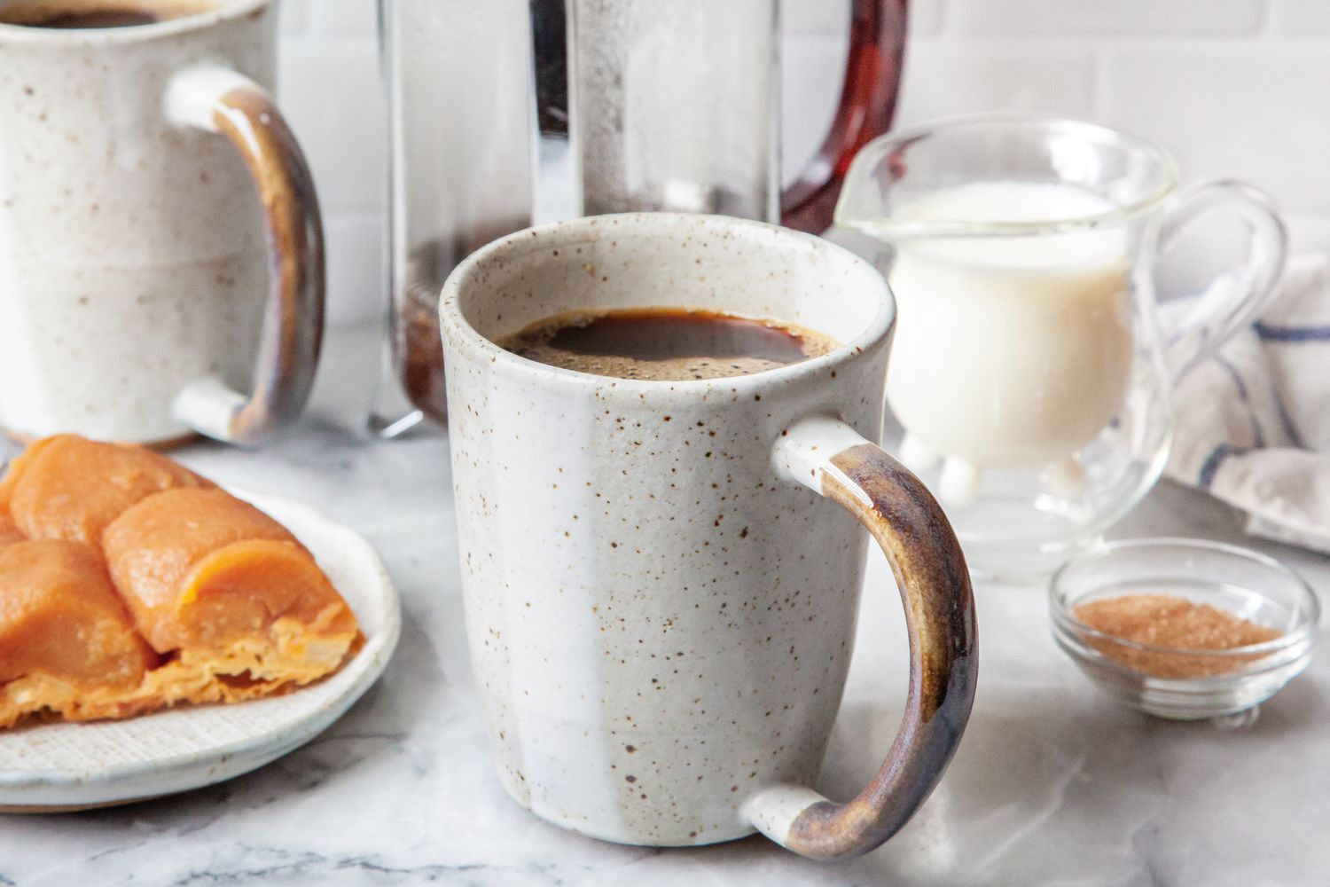 Cup of French Press Coffee Surrounded by a Plate with a Breakfast Pastry, a Bowl of Sugar, a Small Pitcher of Milk, Another Cup of Coffee, and a French Press