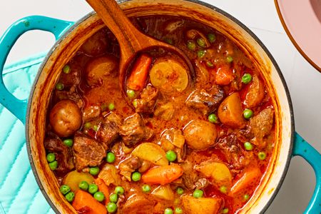 Overhead closeup view of hands-off beef stew in a teal colored dutch oven with a wooden spoon, all on a tile countertop next to a pink bowl and blue hotpad