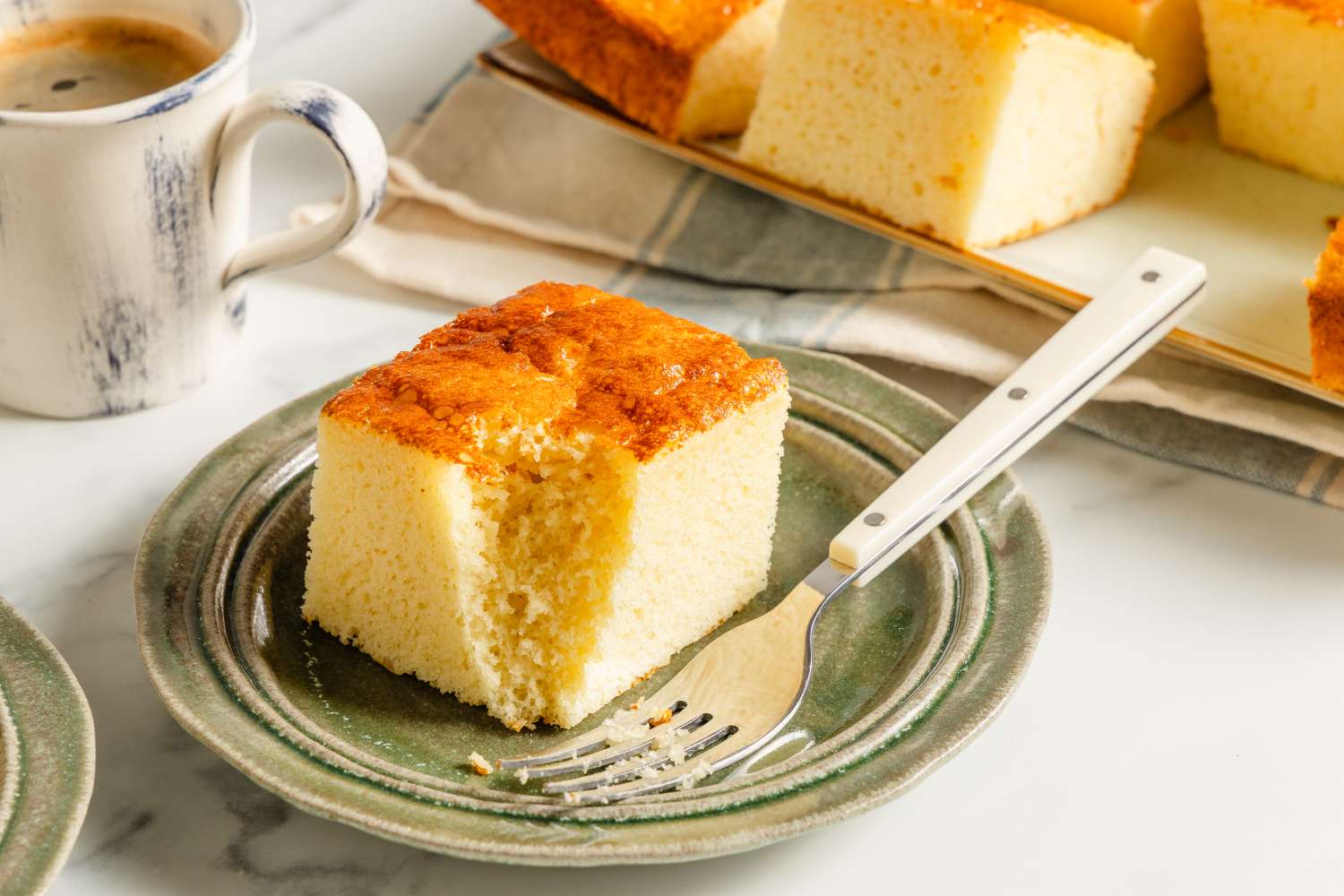 A slice of cake on a plate with a fork, with a cup of coffee and more cake visible in the background