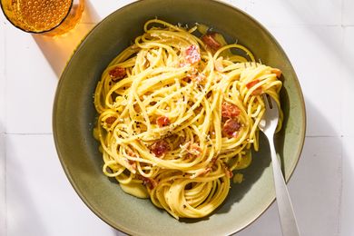 A plate of pasta carbonara with grated cheese and bits of meat with a fork and a glass of beverage nearby