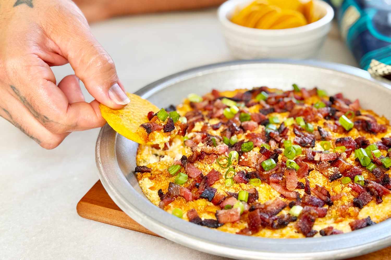 Hand dipping a chip into a bacon and cheese topped dip in a metal dish