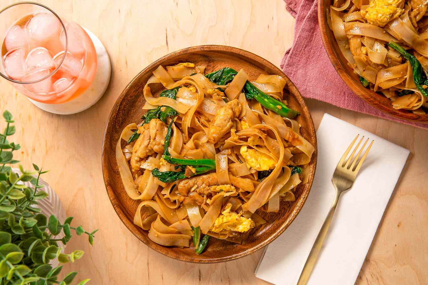 plate of pad see ew at a table setting with another plate of pad see ew, an iced drink, and a table plant