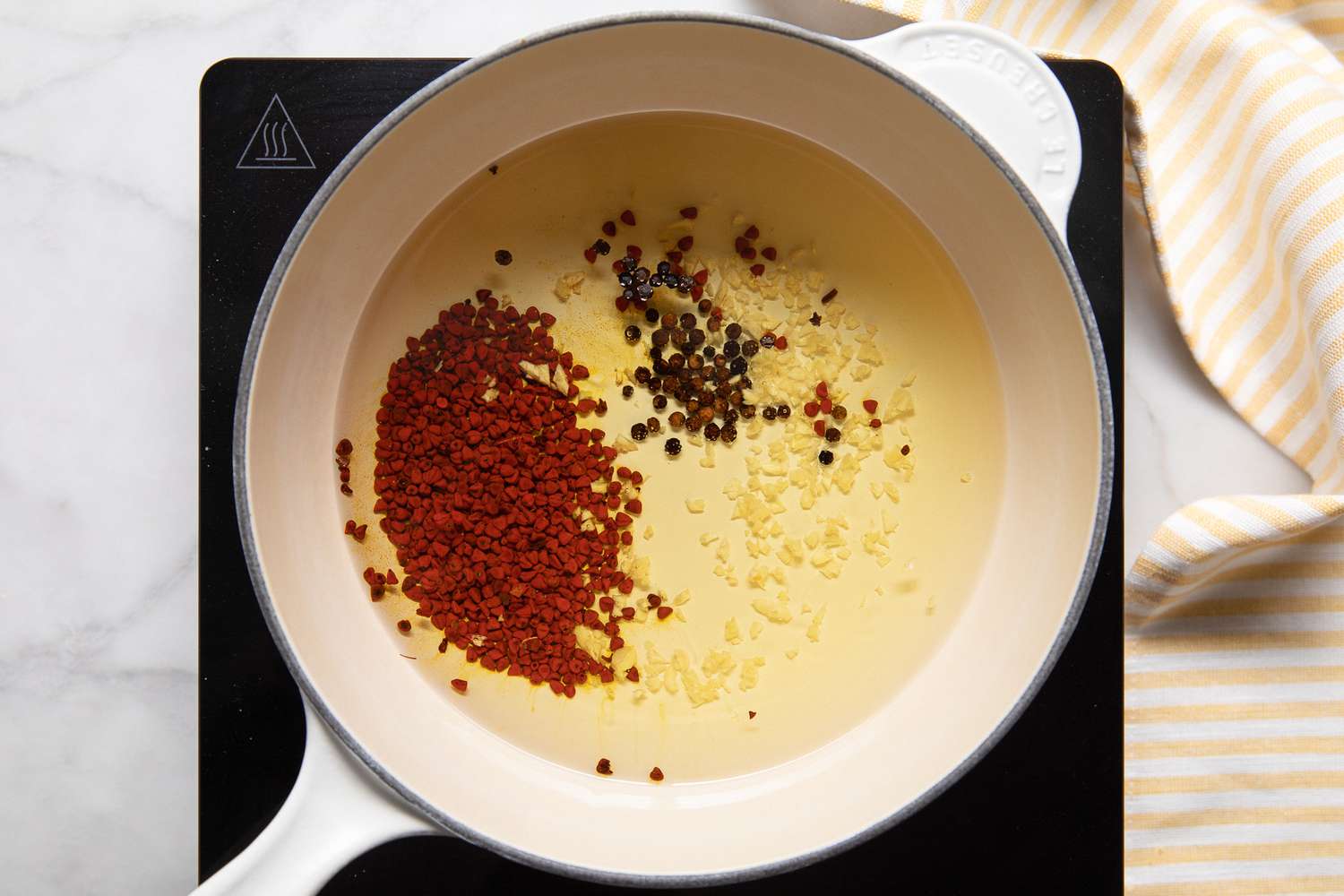 Annatto Seeds, Peppercorns, Garlic, and Vegetable Oil in a Pot Over a Portable Burner, and on the Counter Next to It, a Yellow and White Striped Kitchen Towel for Banana Ketchup Recipe