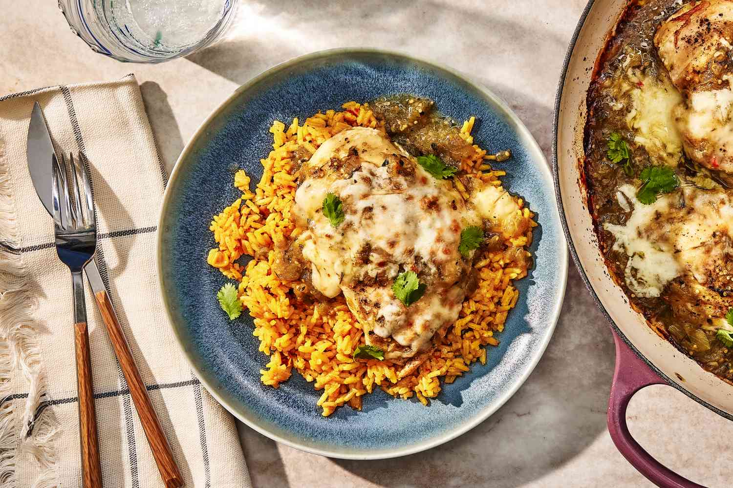 plate of salsa verde chicken over a bed of rice at a table setting with more chicken in a dutch oven, utensils on a table napkin, and a glass of water