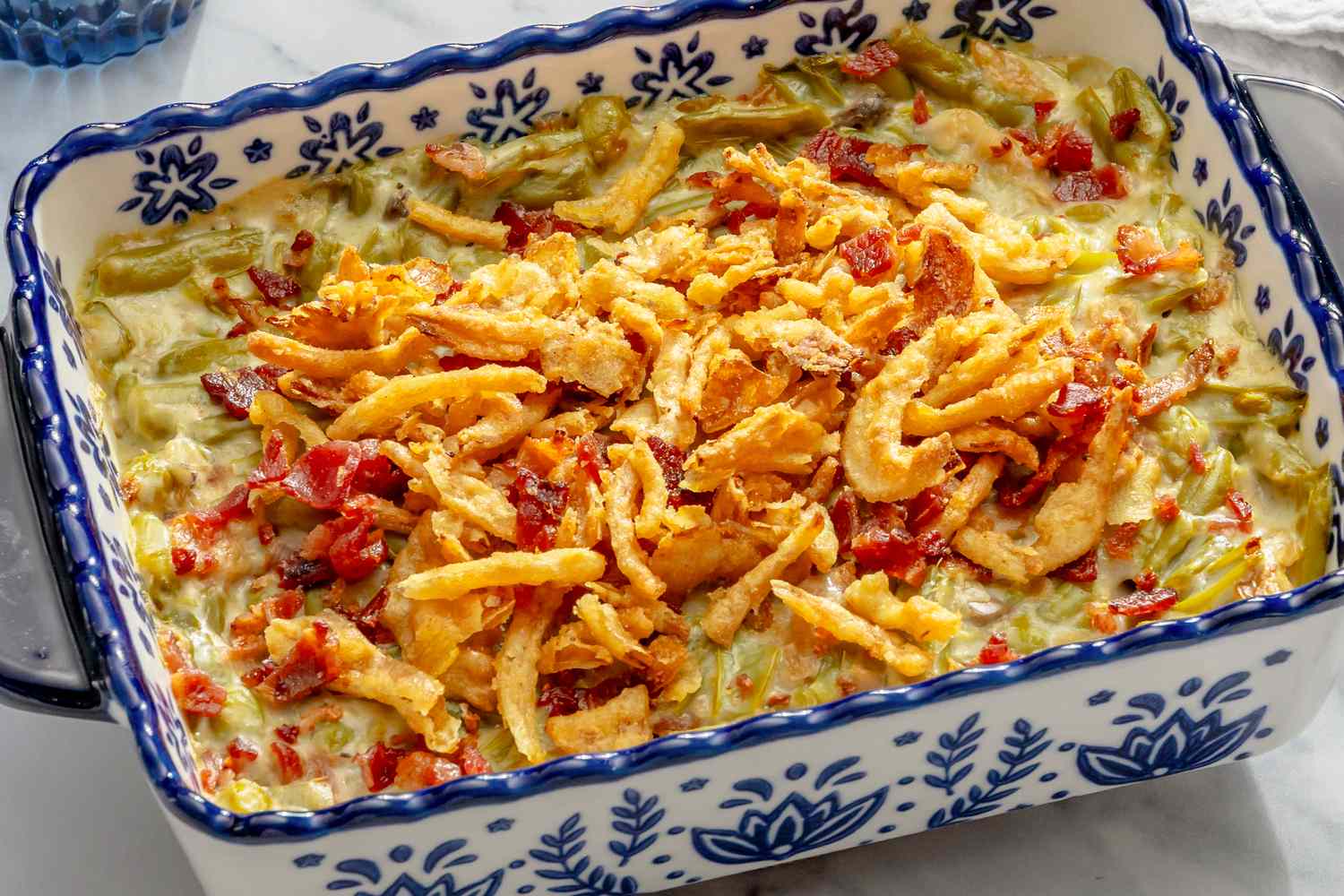 Green bean casserole in a decorative baking dish topped with crispy fried onions and red peppers