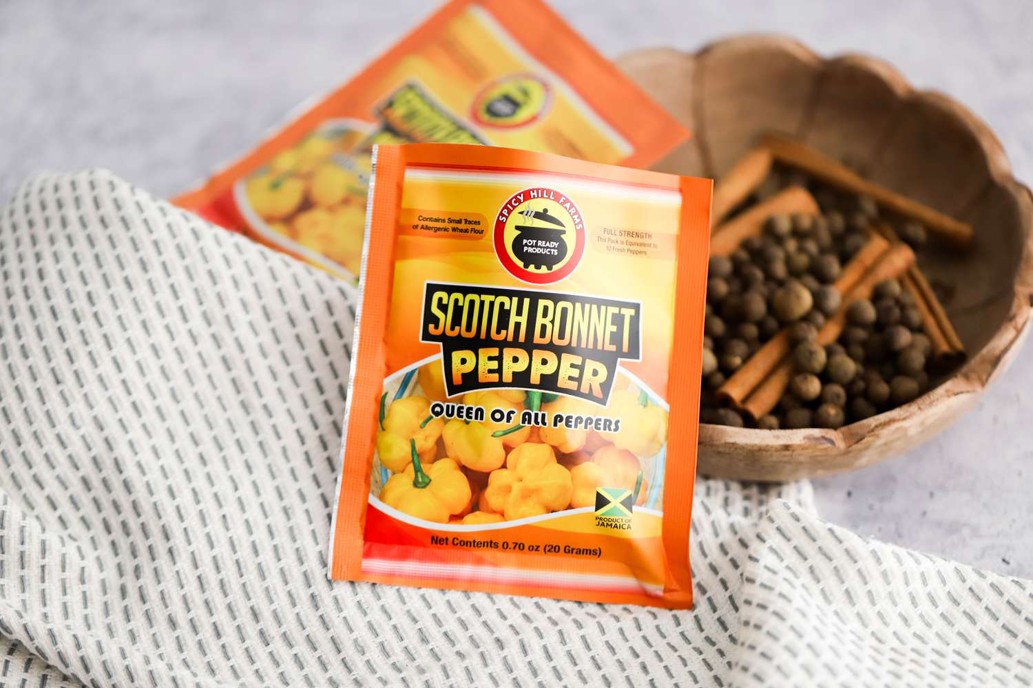 Small Packet of Scotch Bonnet Pepper and in the Surroundings, a Bowl of Whole Spices, Another Packet of Dried, Ground Scotch Bonnet Peppers, and a Kitchen Towel