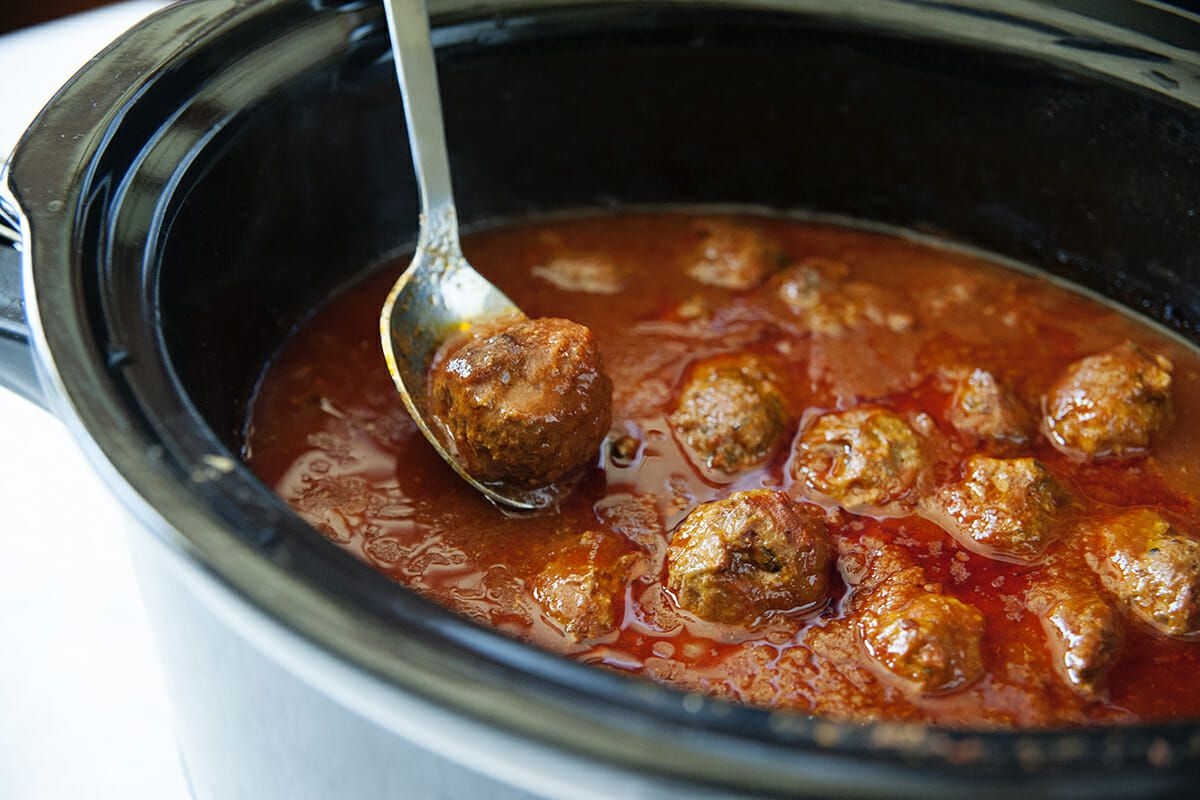 Meatballs in a slow cooker filled with tomato sauce with a spoon lifting a meatball out of the sauce.