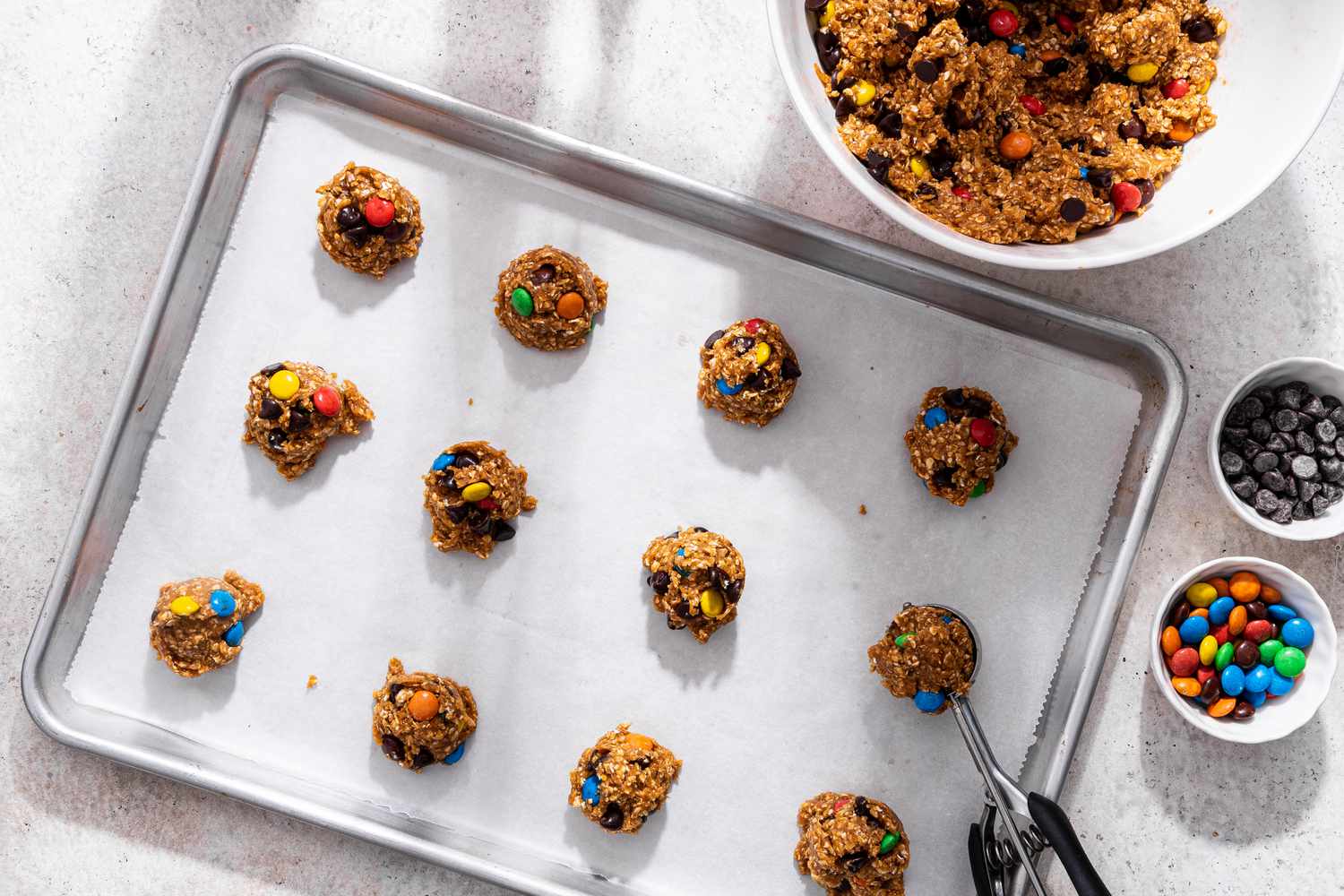 Dollops of monster cookie dough added to a parchment paper lined baking sheet using a cookie scoop, and in the surroundings, the bowl with more monster cookie dough and two small bowls with more mix-ins (one with chocolate chips, the other with M&M's)