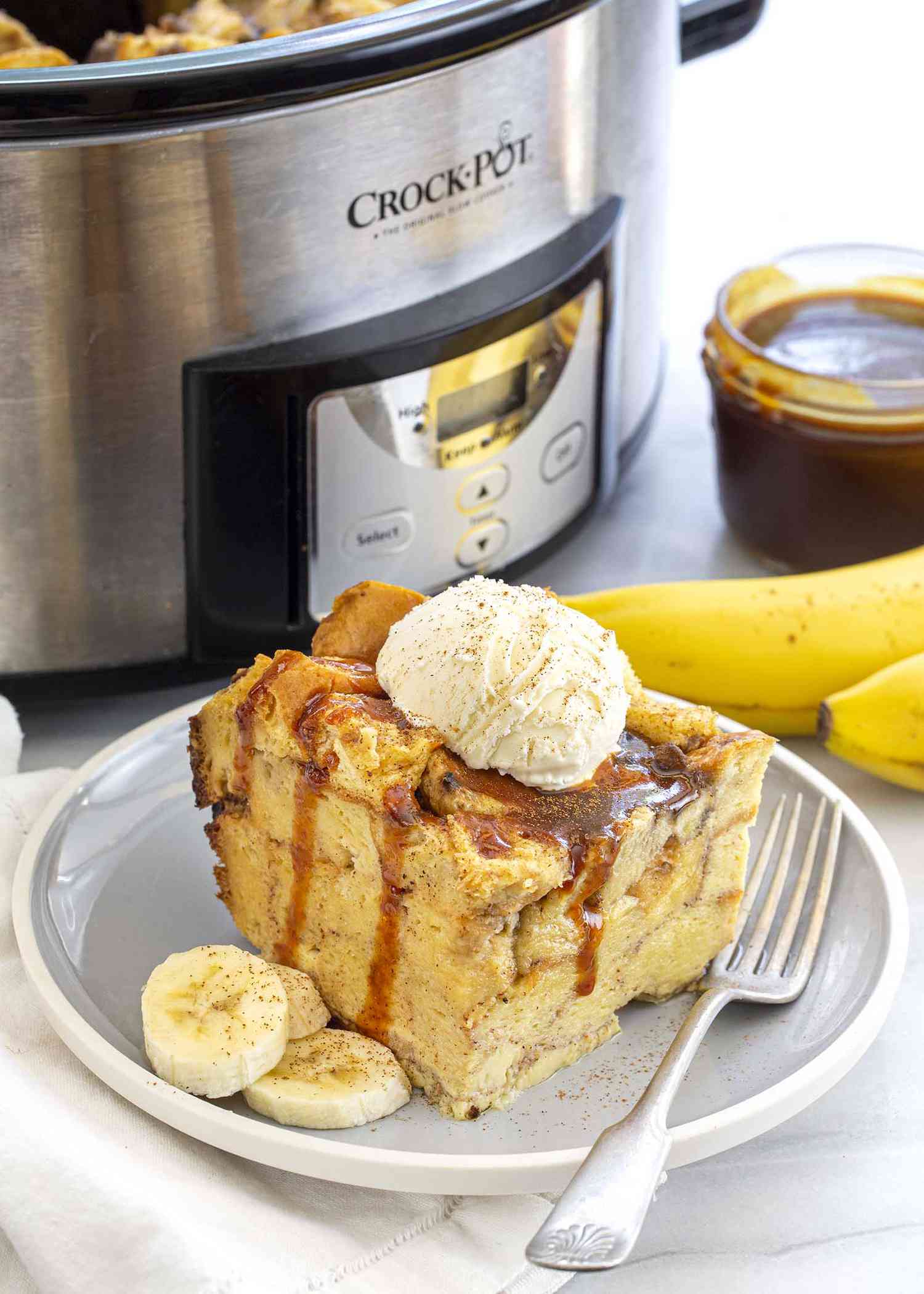 Slice of easy to make slow cooker banana bread pudding topped with a scoop of ice cream and drizzeld with caramel sauce on a gray plate with a fork and sliced bananas. The slow cooker and a jar of caramel sauce is off ot the side.