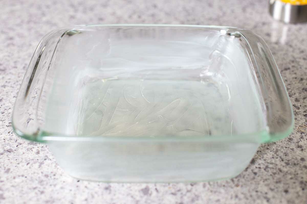 A square glass casserole dish is buttered to make Cheesy Cauliflower.