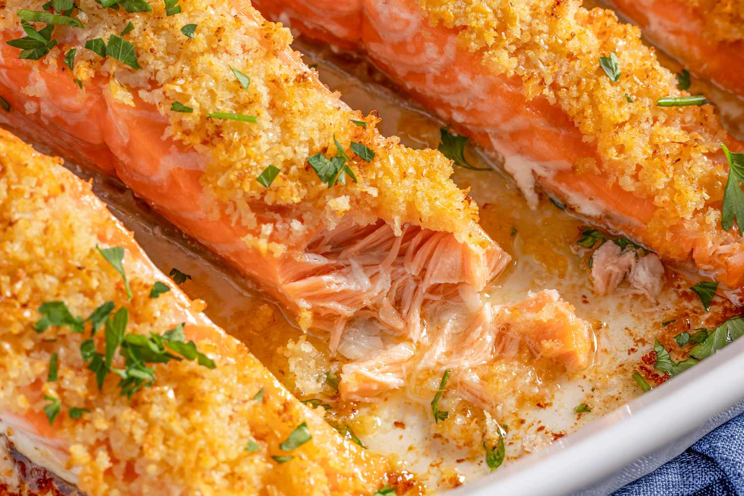 Closeup view of a white baking dish of four salmon fillets topped with garlic butter, panko, and parmesan mixture and sprinkled with parsley