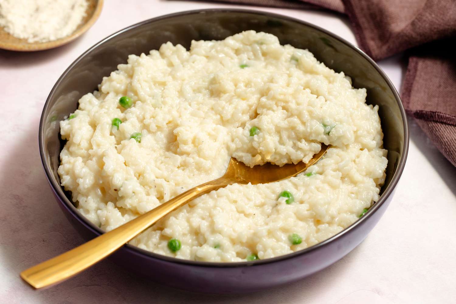 Closeup view of a brown bowl of risotto with peas and a gold spoon on a pink tinted countertop next to a brown table napkin